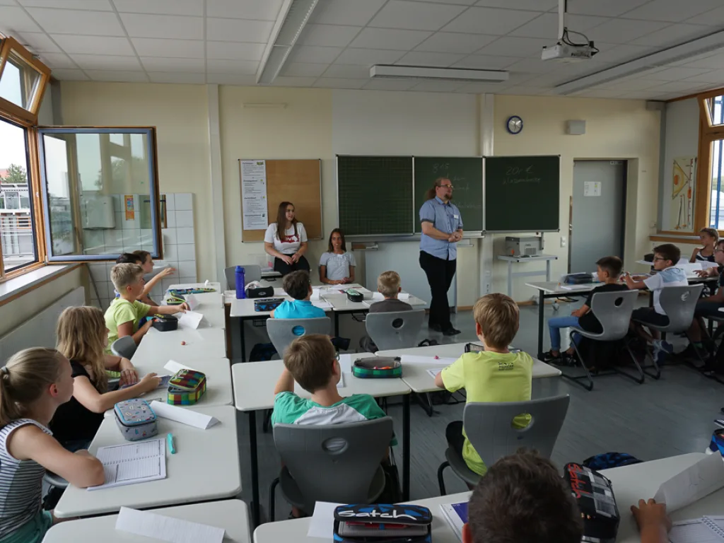 Lehrkraft steht vor der Tafel, Kinder hören zu.